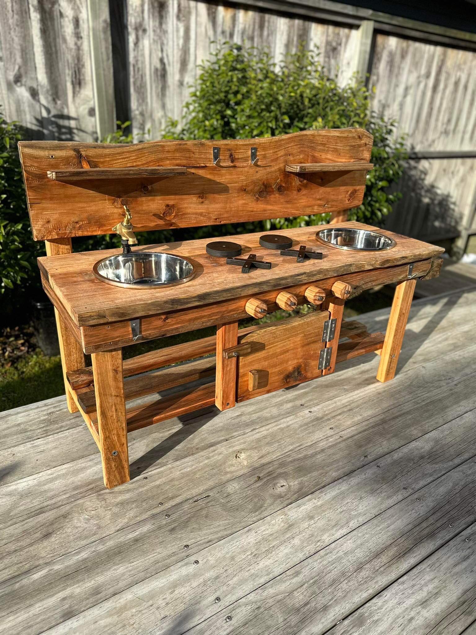 Outdoor Mud play kitchen for children and families in backyards. Made with Macrocarpa wood. It has a double sink and a working outdoor tap to connect to a hose. Stainless steel hooks and hinges. Opening oven door and hotplates for pretend play cooking. 