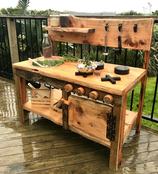 High quality Macrocarpa outdoor mud kitchen for children handmade in New Zealand. Natural Macrocarpa wood and oiled to finish. Includes a stainless steel sink bowl, four hobs, four turning knobs, 3 hooks and a floating shelf. one metre long. 